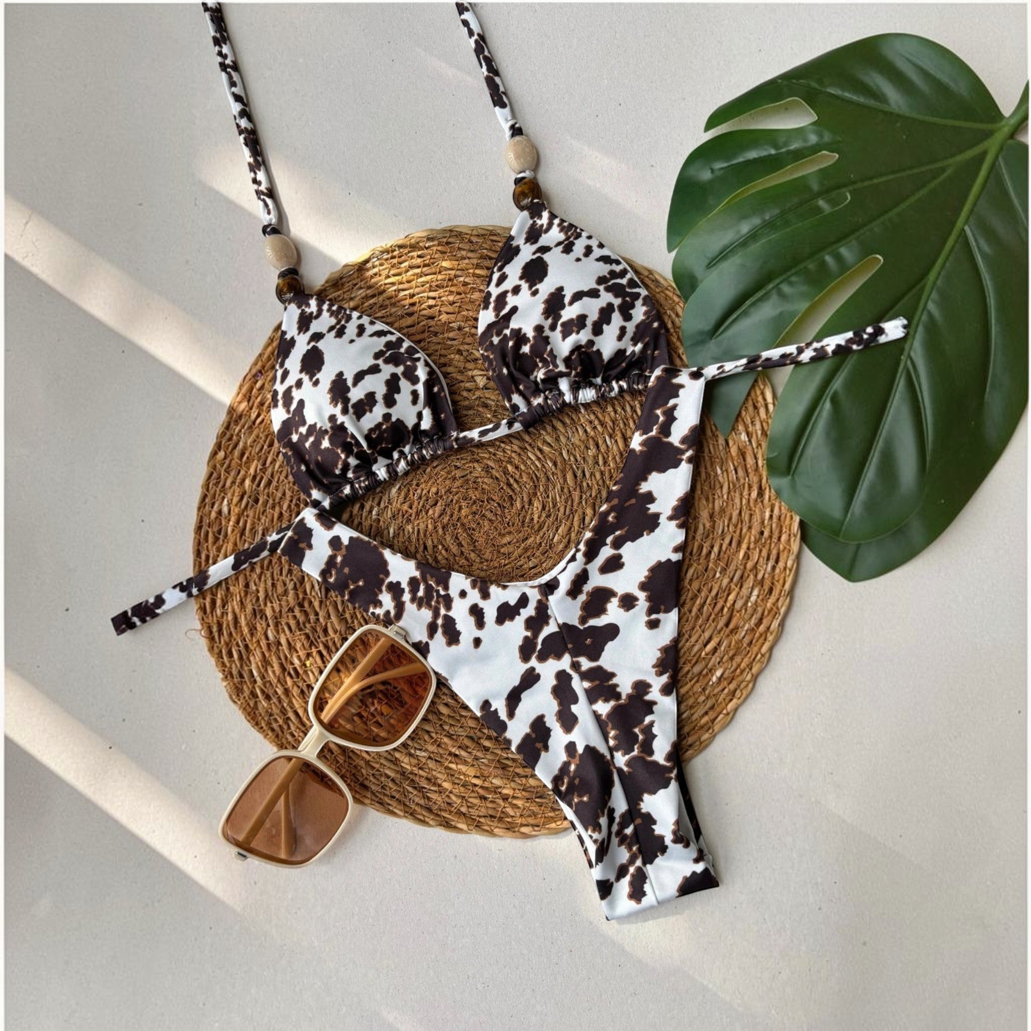 Biquíni GRINE modelo cortininha e asa delta com estampa cow print, sobre base de palha com óculos de sol.