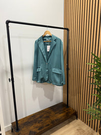 Blazer oversize acetinado verde GRINE com um botão e bolsos frontais, pendurado em cabide num expositor de loja