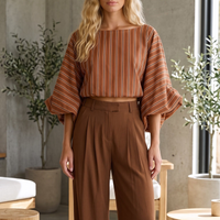Modelo loira veste blusa curta GRINE listrada em tons terrosos com mangas bufantes volumosas e decote canoa