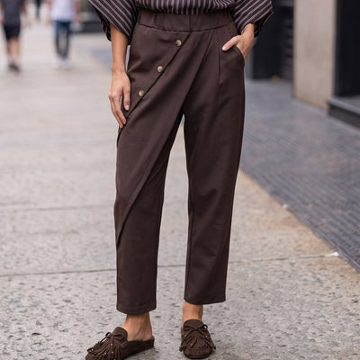 Calça envelope GRINE castanha com fecho diagonal, botões prateados e corte fluido em ambiente urbano.