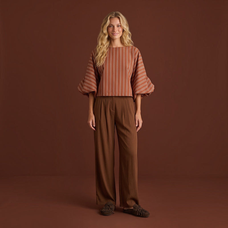 Modelo loira veste calças pantalona fluidas castanhas da GRINE com blusa às riscas e sapatos confortáveis.