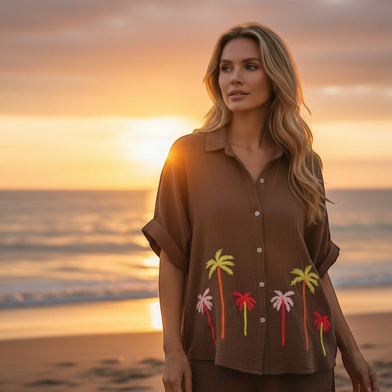 GRINE camisa de algodão castanha com bordados de palmeiras coloridas, corte oversize, usada por uma mulher na praia ao pôr do sol.