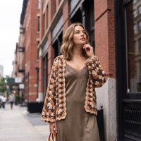 Modelo veste casaco de croché hexagonal GRINE castanho de corte curto sobre vestido acetinado em ambiente urbano