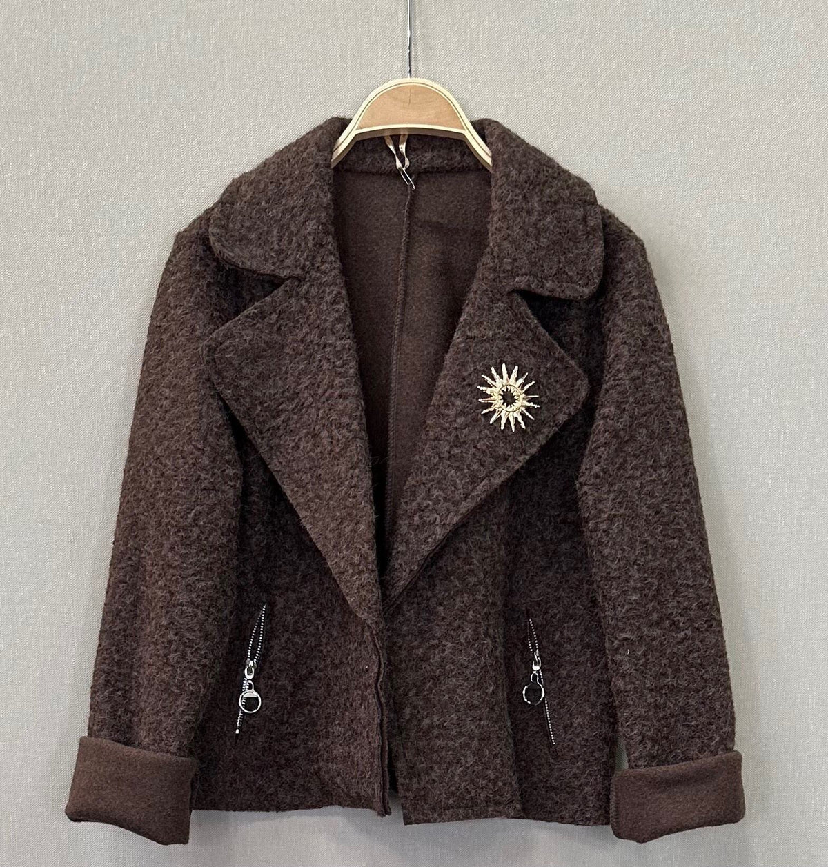 Casaco curto castanho GRINE com textura macia, dois bolsos falsos com fecho e pregadeira decorativa na gola.
