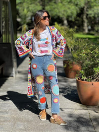 Casaco/kimono comprido de crochet GRINE com padrão colorido e ponto aberto, usado por uma mulher com calças de ganga decoradas com aplicações de crochet.