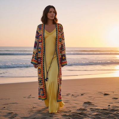 GRINE Casaco/kimono comprido de crochet com padrão colorido e ponto aberto, usado sobre vestido amarelo na praia ao pôr do sol.