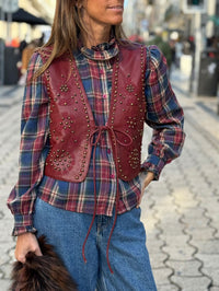 Colete GRINE Chester em PU bordeaux com bordados e tachas, fechado com atilho frontal sobre camisa aos quadrados.