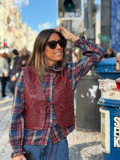 Mulher usa colete GRINE Chester em PU bordeaux com bordados e tachas sobre camisa de xadrez em ambiente urbano