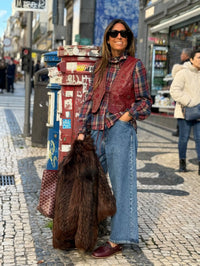 Mulher usa colete GRINE bordeaux em PU com tachas e bordados, camisa de xadrez e calças de ganga numa rua urbana.