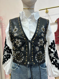 Colete GRINE Chester em PU preto com bordados florais e tachas douradas, fechado com atilho frontal sobre camisa