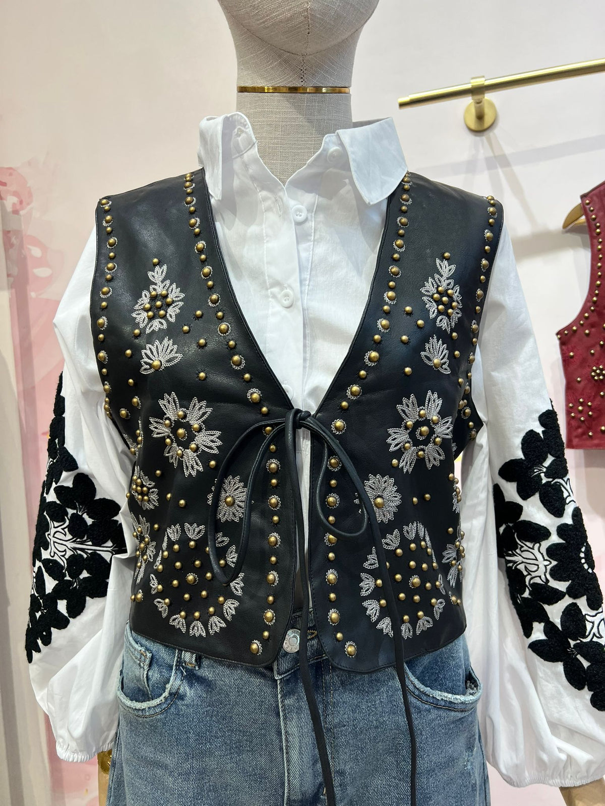 Colete GRINE Chester em PU preto com bordados florais e tachas douradas, fechado com atilho frontal sobre camisa