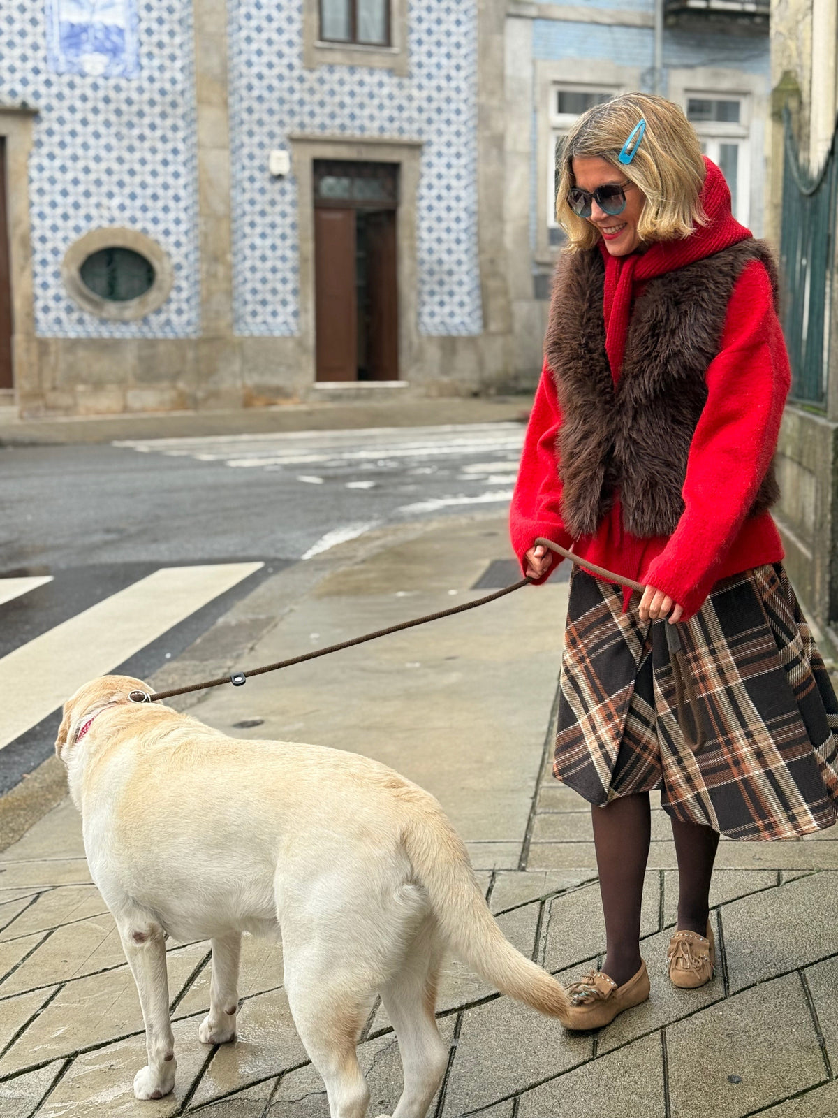 Mulher a passear cão com colete de pêlo castanho GRINE sobre camisola vermelha e saia xadrez em ambiente urbano