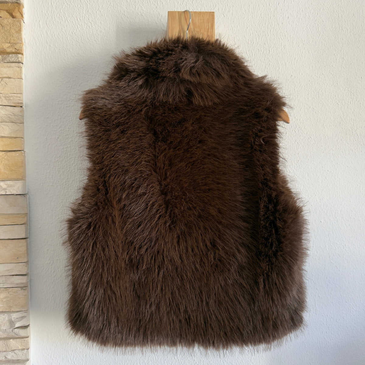 Colete de pêlo castanho GRINE, elegante e quente, pendurado num cabide de madeira contra uma parede branca.