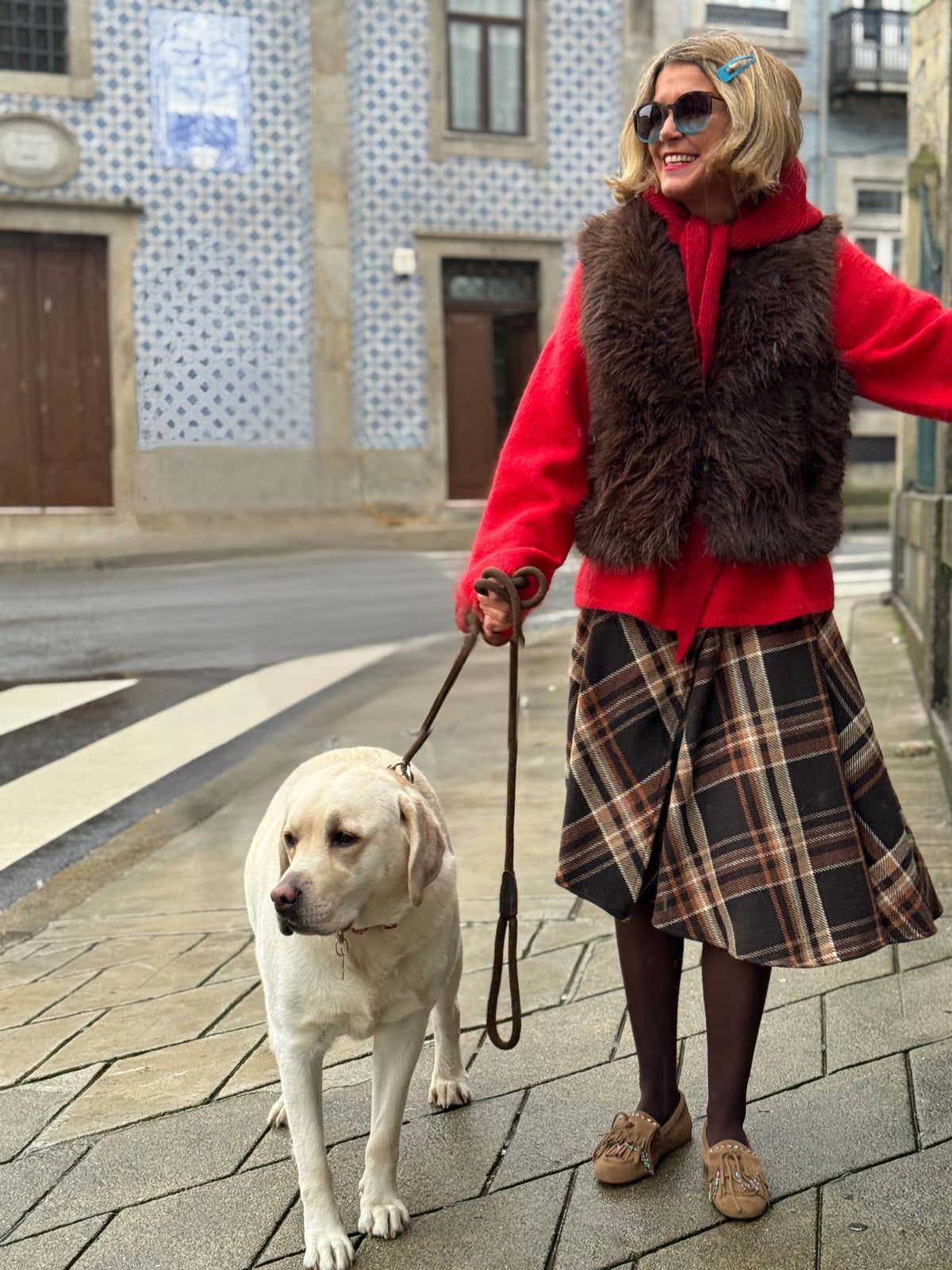 Mulher a passear um cão labrador usando colete de pêlo castanho GRINE, camisola vermelha e saia xadrez na rua.