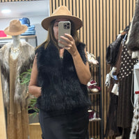 Mulher a usar colete de pêlo preto elegante da GRINE com chapéu castanho em frente ao espelho