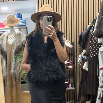 Mulher a usar colete de pêlo preto elegante da GRINE com chapéu castanho em frente ao espelho