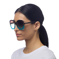 Modelo usa óculos de sol GRINE OKKIA ANNA com armação tartaruga e azul e lentes degradé em fundo branco