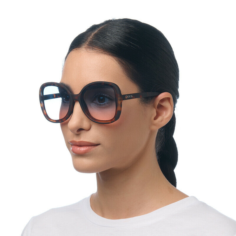 Modelo usa óculos de sol GRINE OKKIA MONICA com armação redonda oversized em padrão Havana e lentes degradé azul rosa