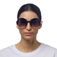 Mulher a usar óculos de sol redondos GRINE OKKIA Monica com armação preta e lentes degradé rosa e pretas