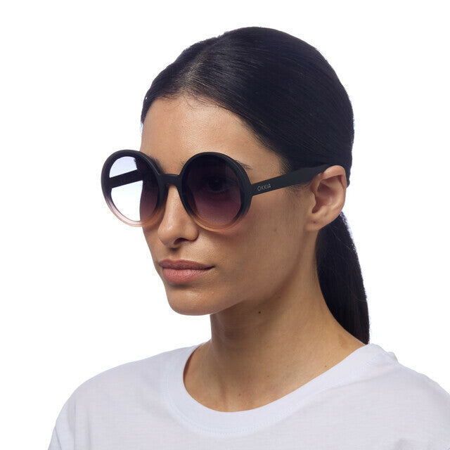 Mulher a usar óculos de sol redondos GRINE OKKIA Monica com armação preta e lentes degradé rosa e preto