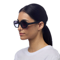 Modelo usa óculos de sol GRINE OKKIA ANDREA pretos com armação hexagonal e lentes em degradé de toque suave