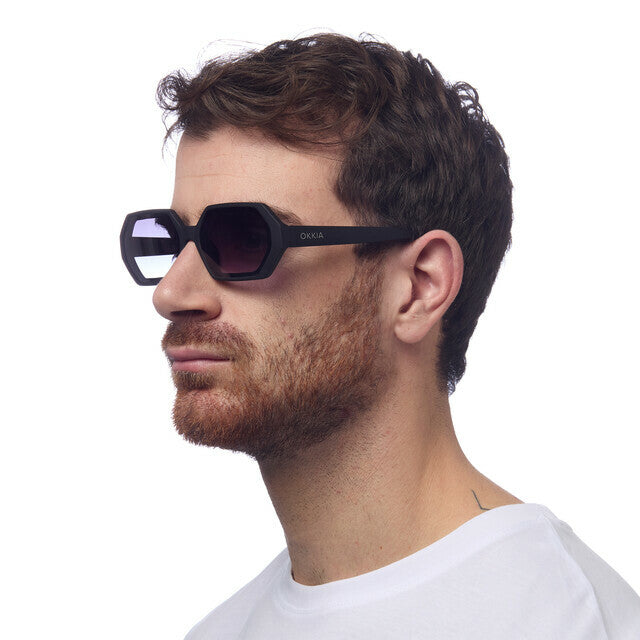 Homem a usar óculos de sol GRINE OKKIA ANDREA com armação hexagonal preta e lentes degradé em fundo branco