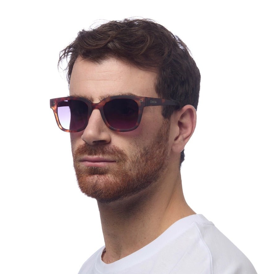Homem a usar óculos de sol GRINE OKKIA Giovanni com armação tartaruga e azul e lentes castanhas polarizadas.