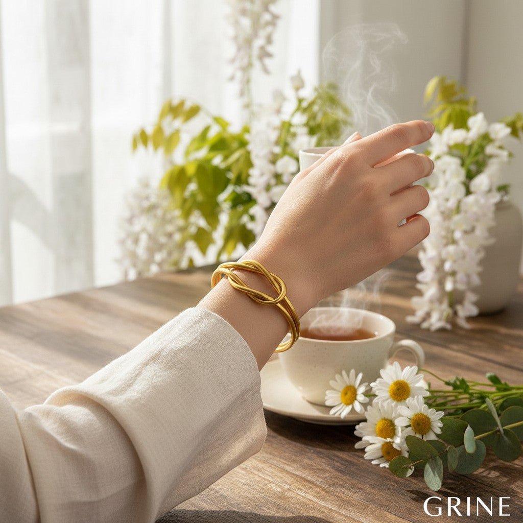 Pulseira Infinity GRINE dourada flexível com design de nó, usada no pulso junto a uma chávena de chá e flores.