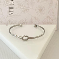 Pulseira GRINE de nó duplo em aço inoxidável prateado com design minimalista e ajustável sobre base branca.