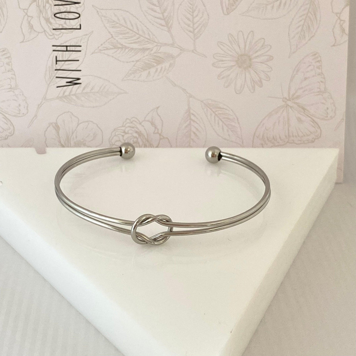 Pulseira GRINE de nó duplo em aço inoxidável prateado com design minimalista e ajustável sobre base branca.