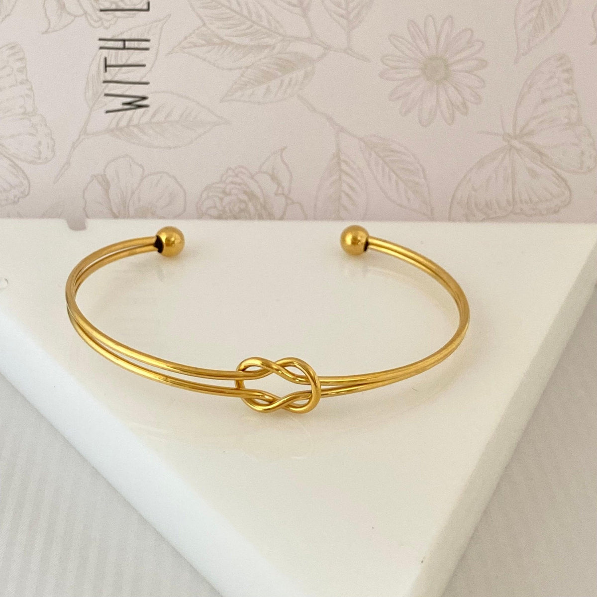 Pulseira GRINE de nó duplo em aço inoxidável dourado com design ajustável e minimalista sobre base branca.