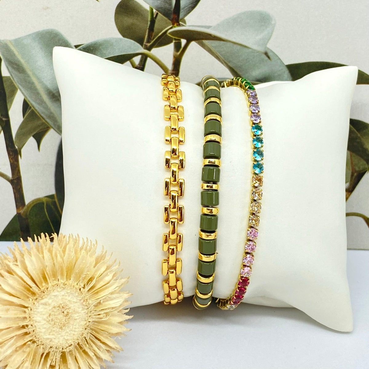 Conjunto de três pulseiras GRINE em aço inoxidável dourado, com pedras de strass coloridas e detalhes em verde.