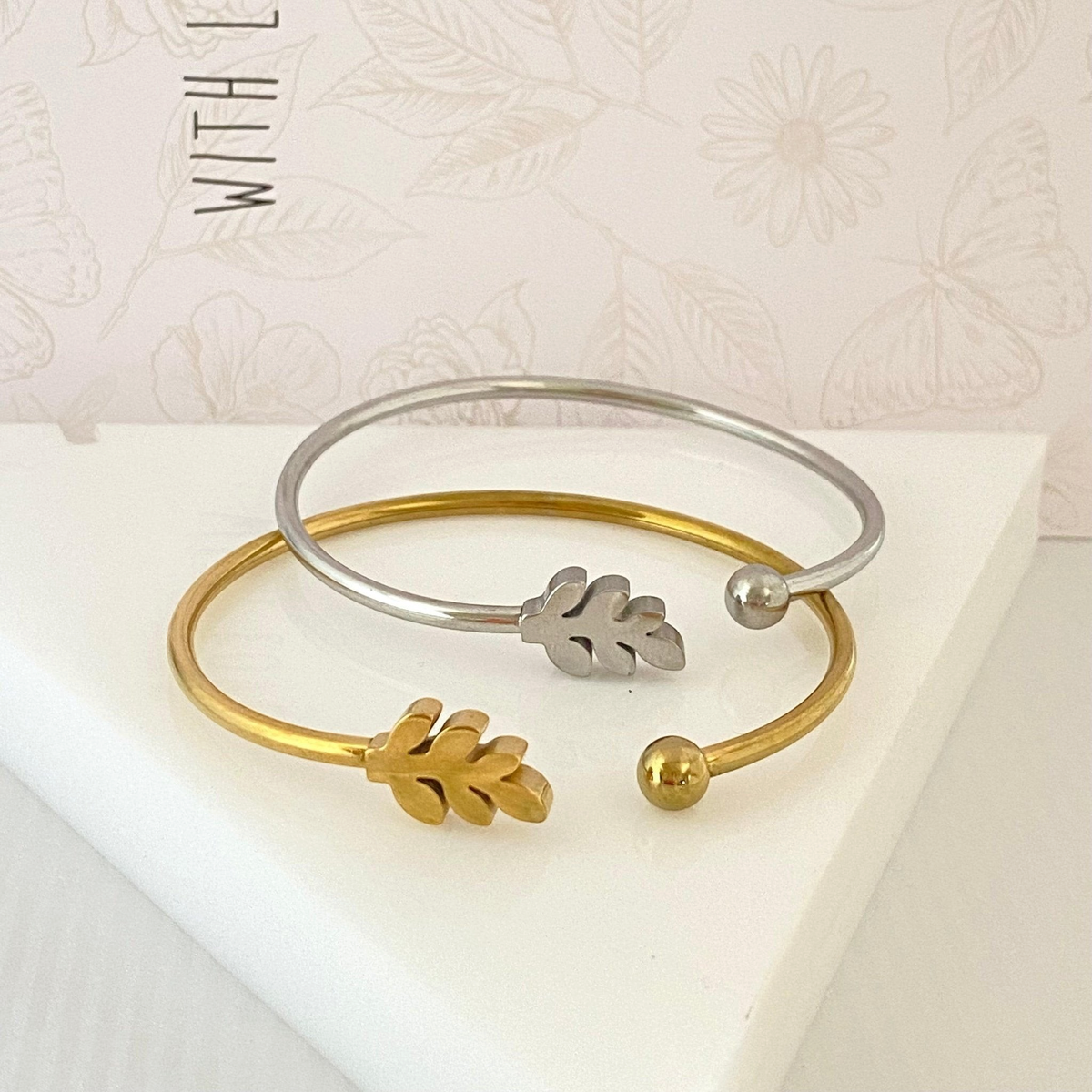 Pulseiras rígidas GRINE em aço inoxidável com detalhe de folha, uma prateada e outra dourada sobre base branca.