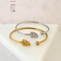 Pulseiras rígidas GRINE em aço inoxidável com detalhe de folha, uma prateada e outra dourada sobre base branca.