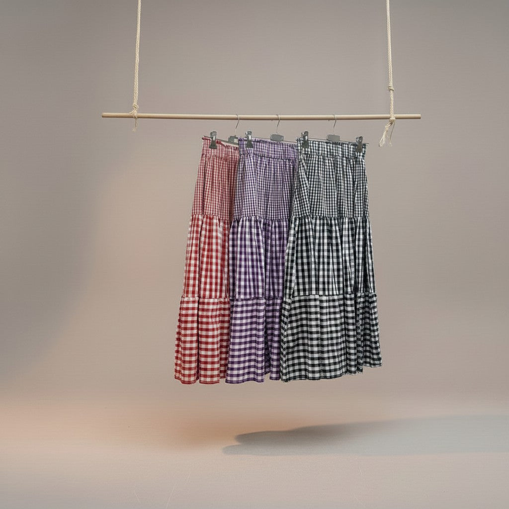 Saia midi GRINE com padrão vichy em camadas, disponível em vermelho, roxo e preto. Cintura elástica e corte fluido.