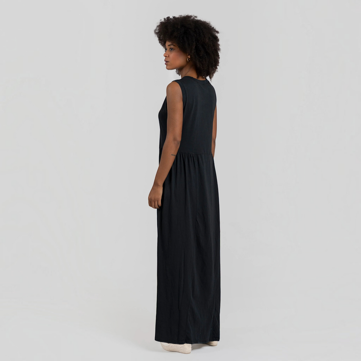 Modelo veste vestido comprido GRINE preto em algodão com corte evasê, decote redondo e sem mangas, vista lateral.